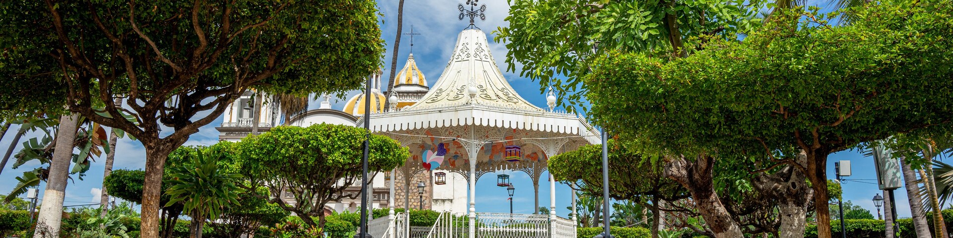Un pabellón con barandillas blancas ornamentadas y un techo abovedado, rodeado de frondosos árboles verdes y un cielo azul despejado.