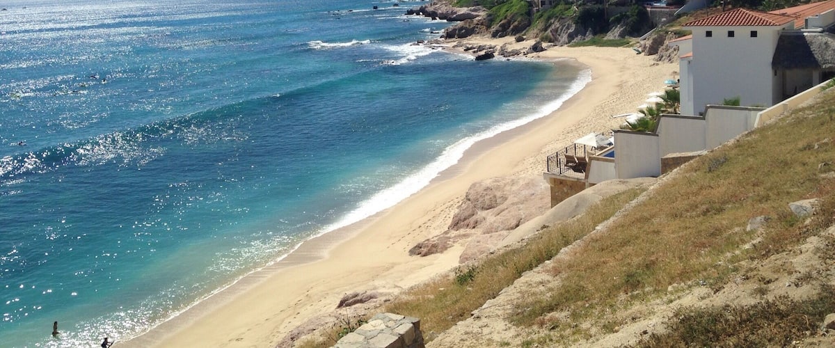A coastal beach with clear blue waters, sandy shore, and a hillside with buildings.