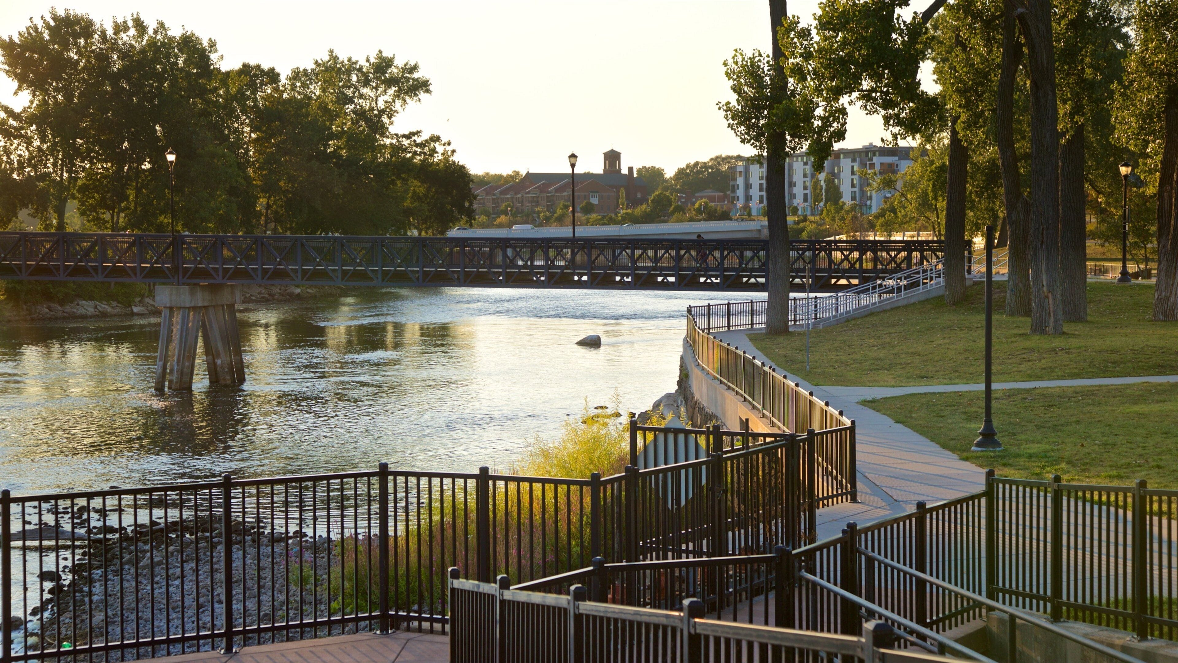 A riverside park with a pedestrian bridge, trees, and buildings in the background.