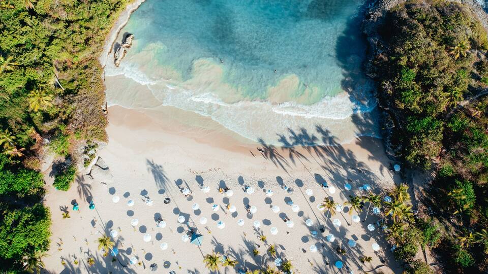 An aerial view of a beach with clear turquoise waters, sandy shore, and numerous umbrellas.