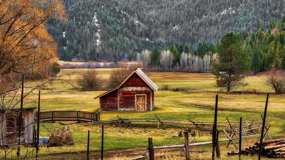A rustic wooden barn in a rural landscape with a forested mountain in the background.
