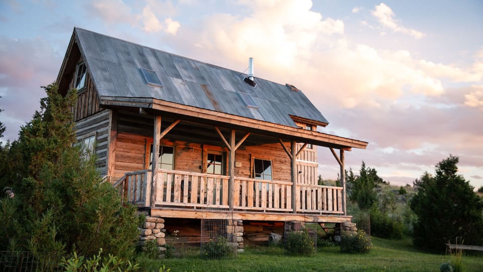 A rustic wooden cabin with a porch, surrounded by greenery and trees.