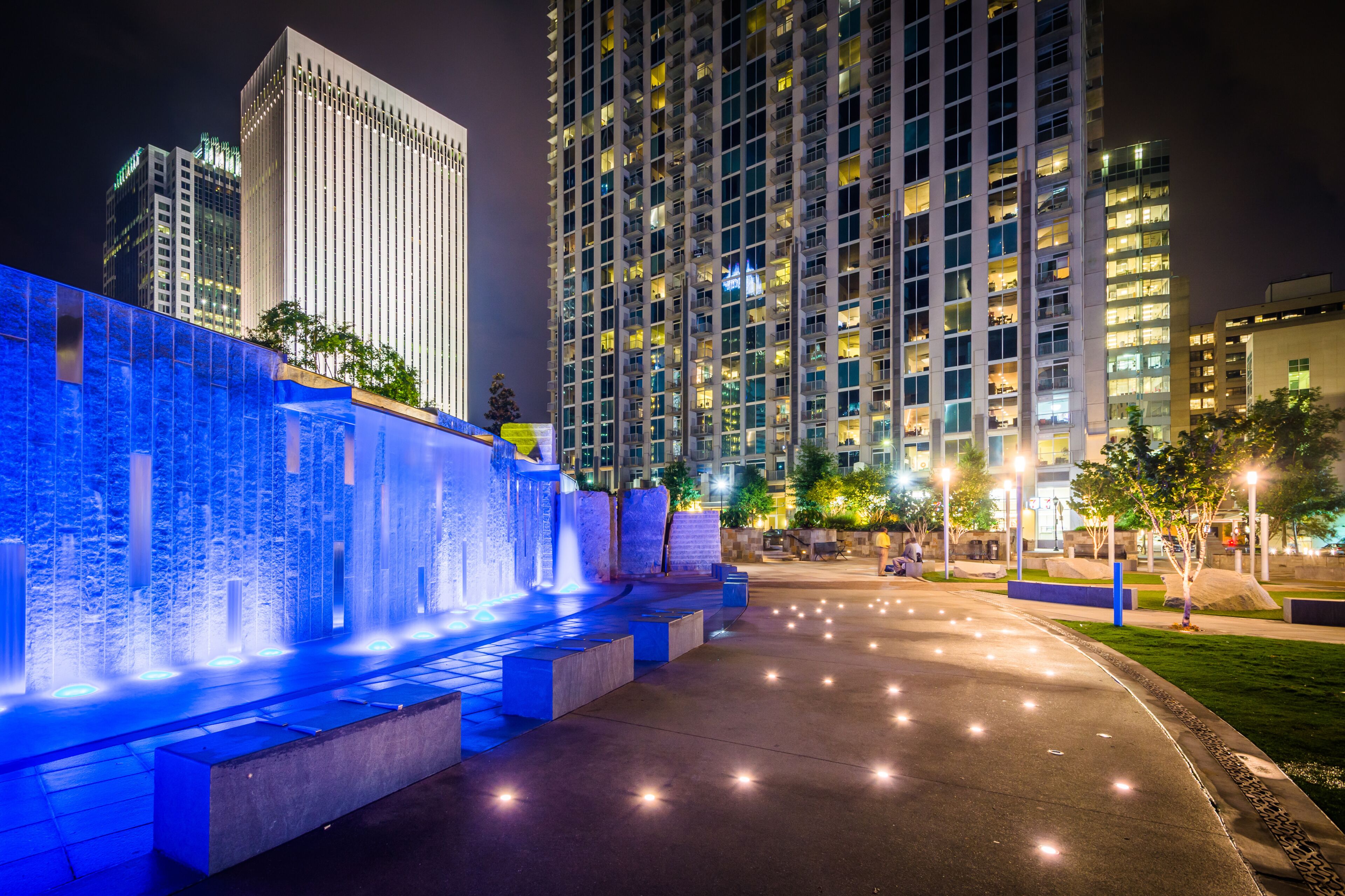 Paisagem urbana noturna com edifícios iluminados, um espelho d'água com iluminação azul e um caminho bem iluminado.