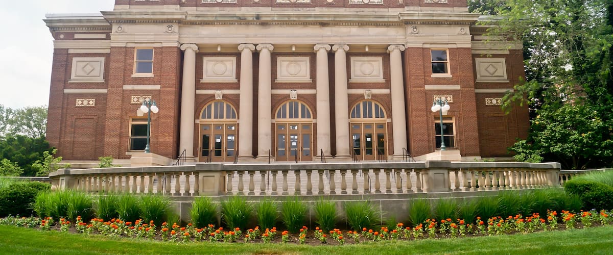 A large brick building with columns and arched windows, surrounded by a garden with flowers and a fountain.