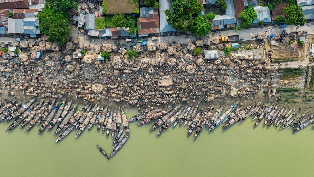 Aerial view of bustling raw jute market with wooden boats on river surrounded by village and greenery, Chinadulli, Islampur, Mymensingh, Bangladesh.