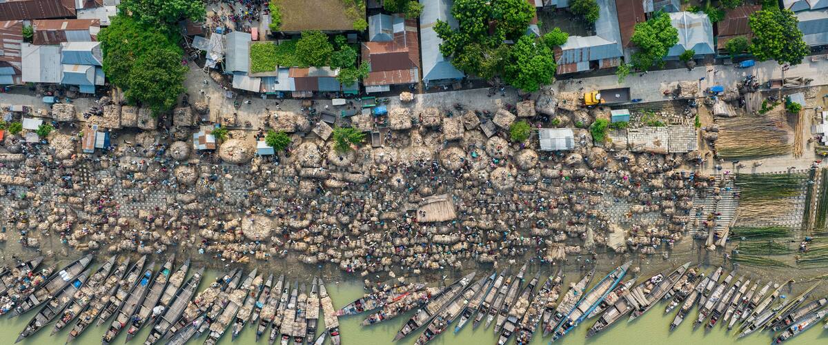 Aerial view of bustling raw jute market with wooden boats on river surrounded by village and greenery, Chinadulli, Islampur, Mymensingh, Bangladesh.