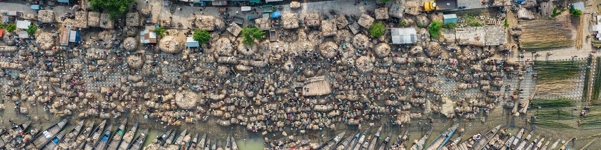 Aerial view of bustling raw jute market with wooden boats on river surrounded by village and greenery, Chinadulli, Islampur, Mymensingh, Bangladesh.