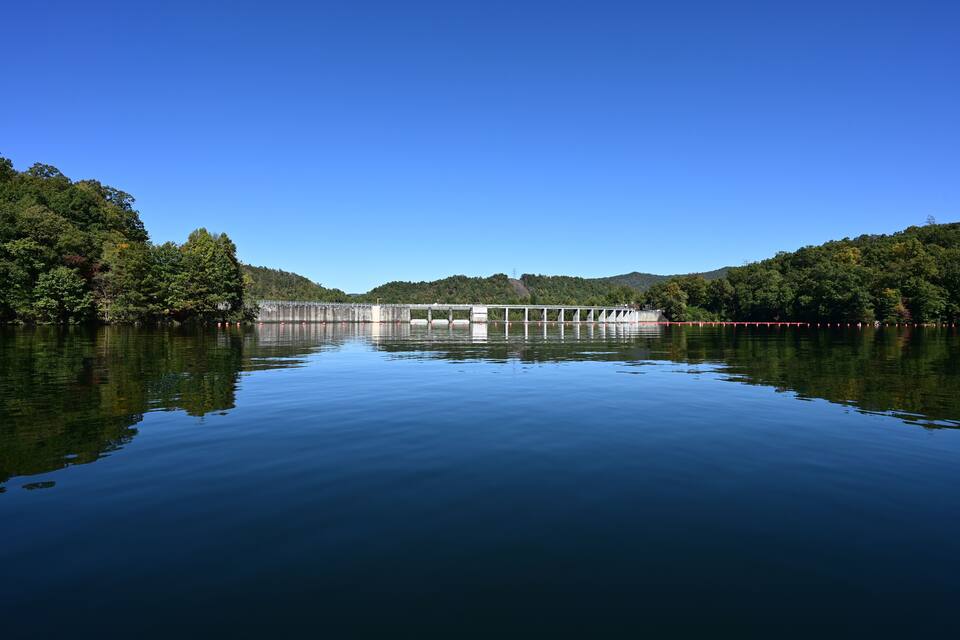 A calm lake with a dam and forested hills in the background.