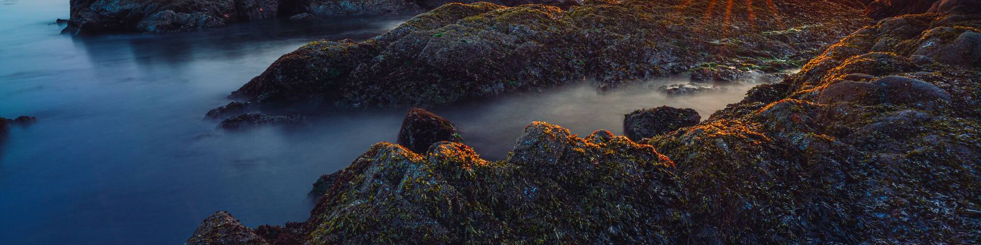 Una costa rocosa al atardecer, con el sol ocultándose tras las rocas.