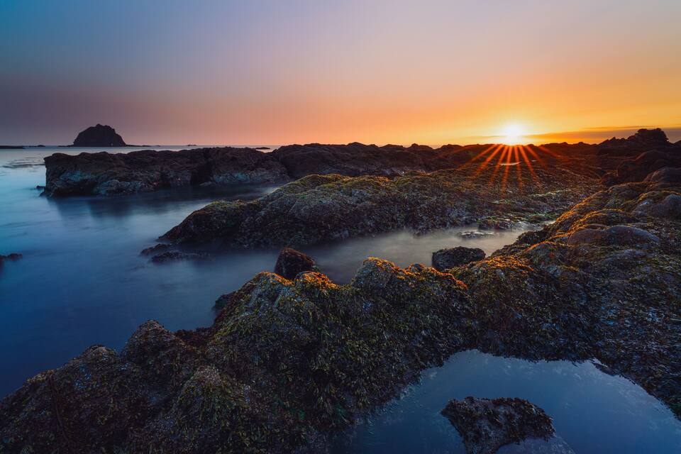 Una costa rocosa al atardecer, con el sol ocultándose tras las rocas.