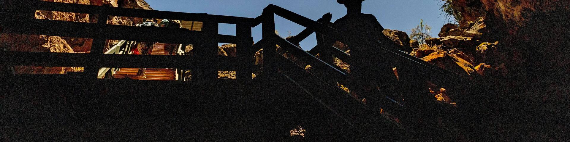 A silhouette of a person wearing a cowboy hat against a clear blue sky, viewed through a rocky archway with wooden stairs.