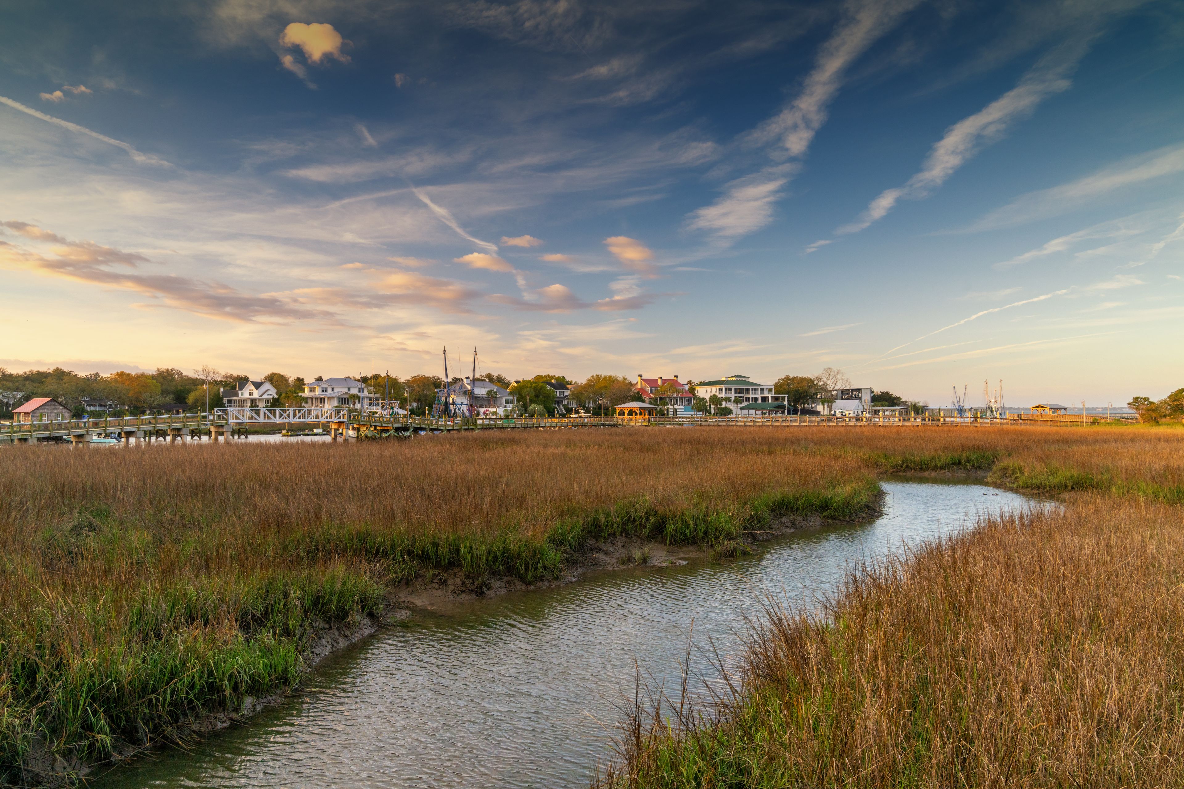 A river meandering through a marshland with houses and a pier in the background.