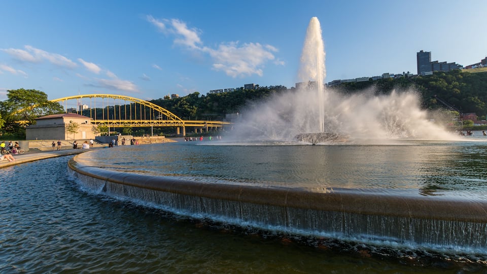 A fountain with water jets, a yellow bridge, and a cityscape in the background.