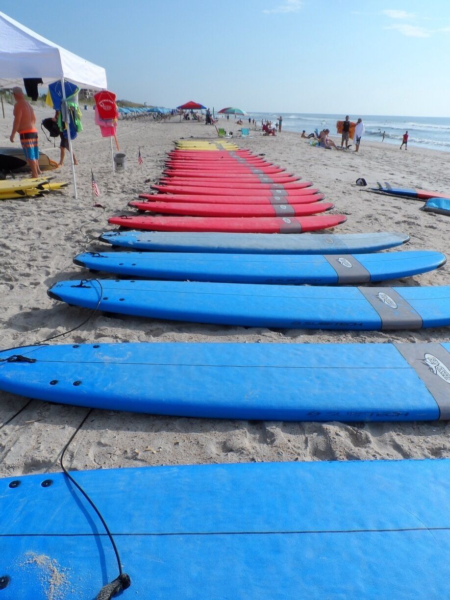 Ein Strand mit reihenweise gestapelten Surfbrettern im Sand, einem Zelt und Personen im Hintergrund.