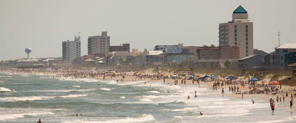 A crowded beach with waves, umbrellas, and buildings in the background.