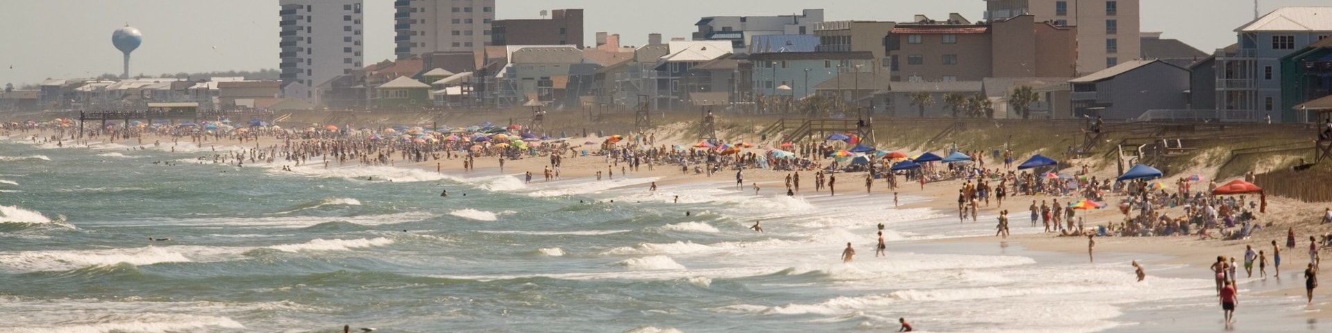 A crowded beach with waves, umbrellas, and buildings in the background.