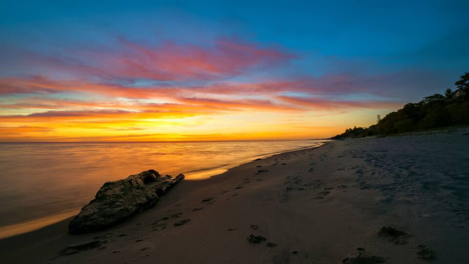 A beach with footprints leading to a large rock, a calm sea, and a sunset with pink and orange clouds.
