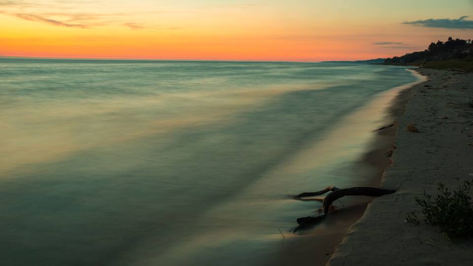 A beach at sunset with calm water and a fallen tree on the sand.