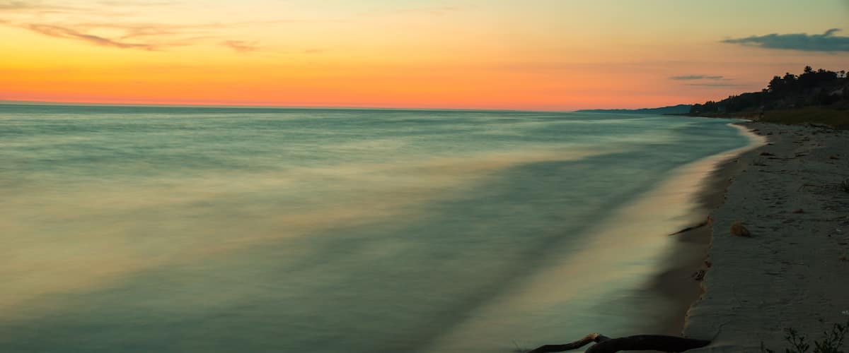 A beach at sunset with calm water and a fallen tree on the sand.
