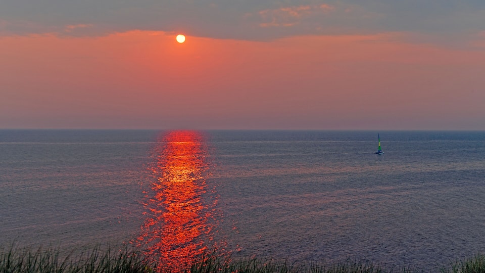 A sunset over a calm sea with a sailboat in the distance.