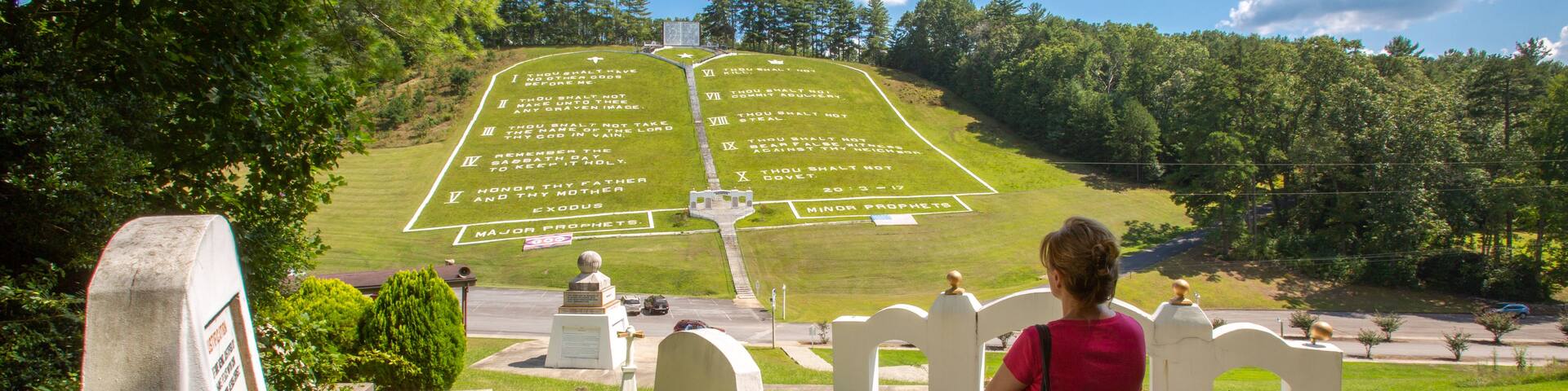A person standing on a set of stairs overlooking a grassy area with a large, grassy cross inscribed with biblical verses.