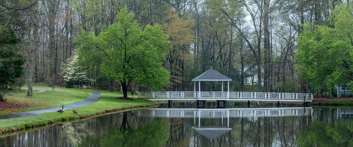 A serene pond with a gazebo, surrounded by lush trees and a walking path.