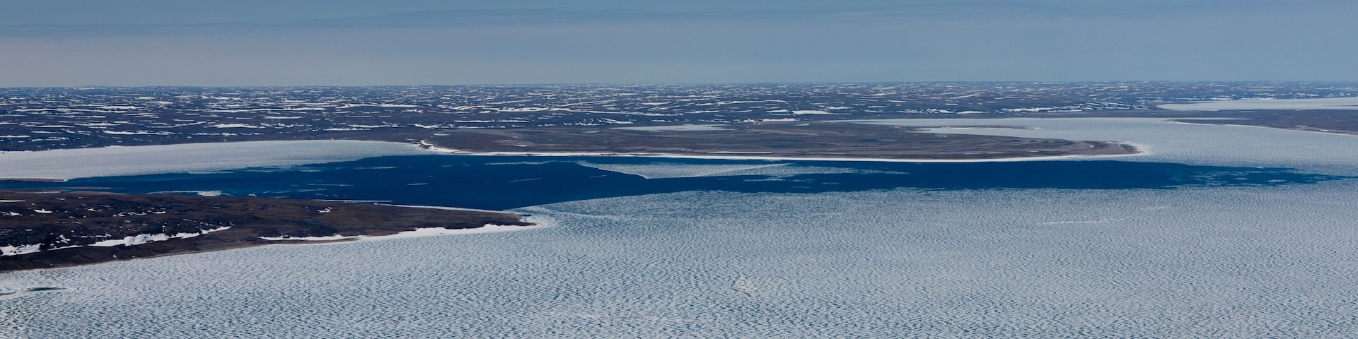 Un lago helado e inmenso rodeado de terreno nevado y edificios al distante.