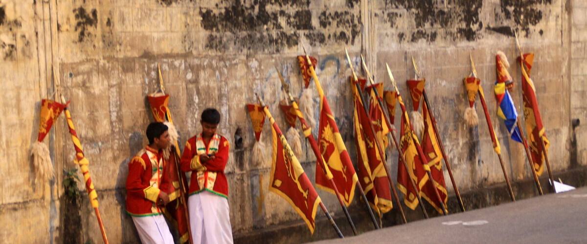 A group of men in traditional attire standing in front of a stone wall with flags.