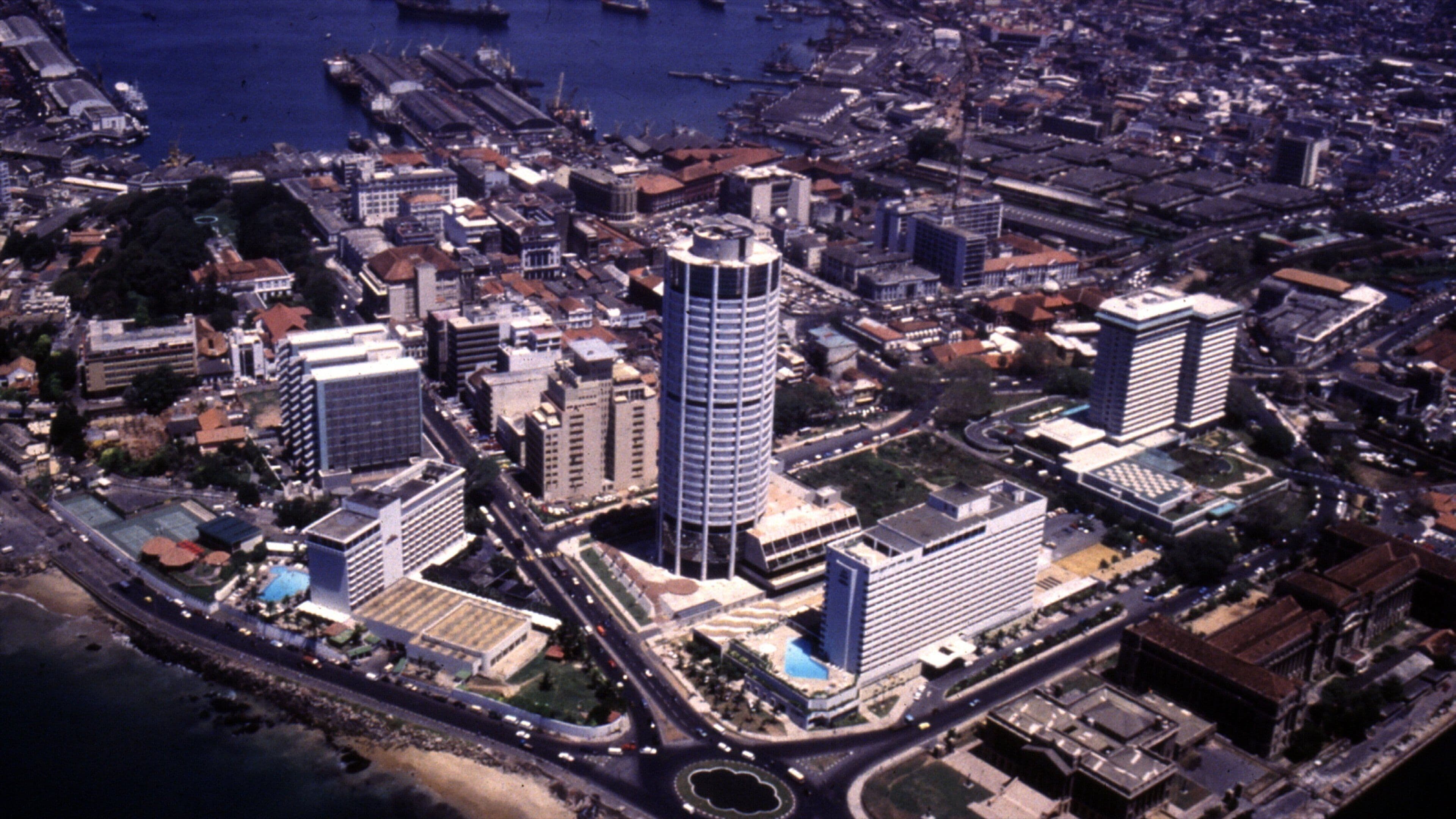 An aerial view of a coastal city with a prominent cylindrical skyscraper, dense urban buildings, and a marina with boats.