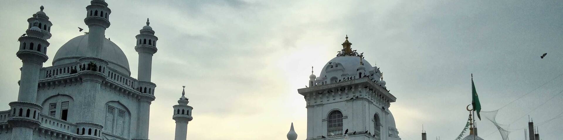 A white mosque with multiple domes and minarets, a sign reading "Shaikh Usman Waliullah Shrine & Masjid," and a bicycle parked nearby.