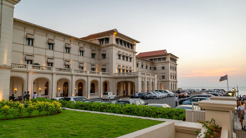 A large, classical building with multiple arches and a red roof, surrounded by a parking lot and a grassy area.