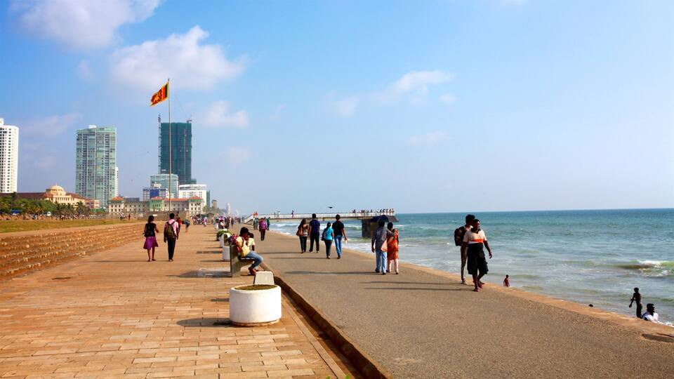 Un paseo marítimo con gente caminando, una bandera y edificios modernos al fondo.