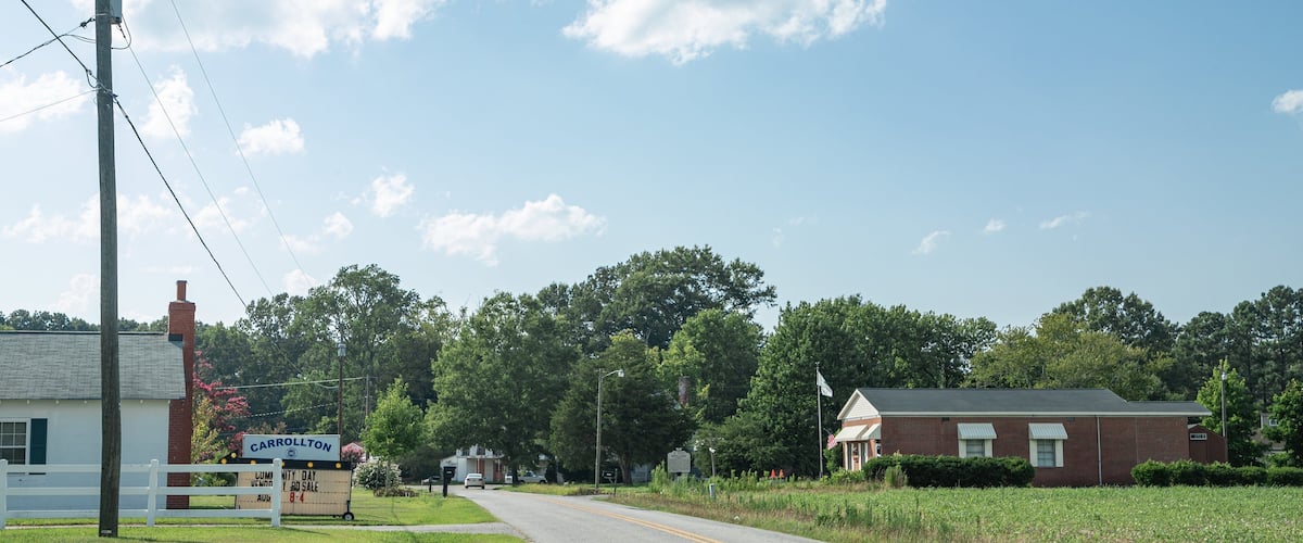 A rural road with a sign for Carrollton, a brick building, and lush greenery.