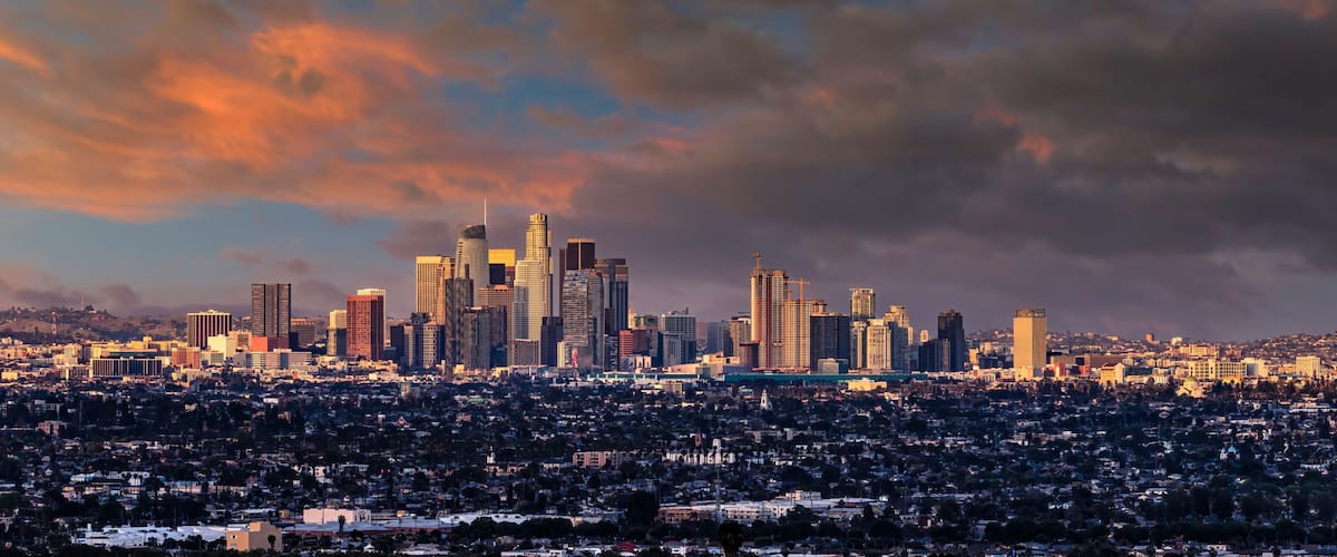 A city skyline with high-rise buildings, a dense residential area in the foreground, and a dramatic sunset sky.