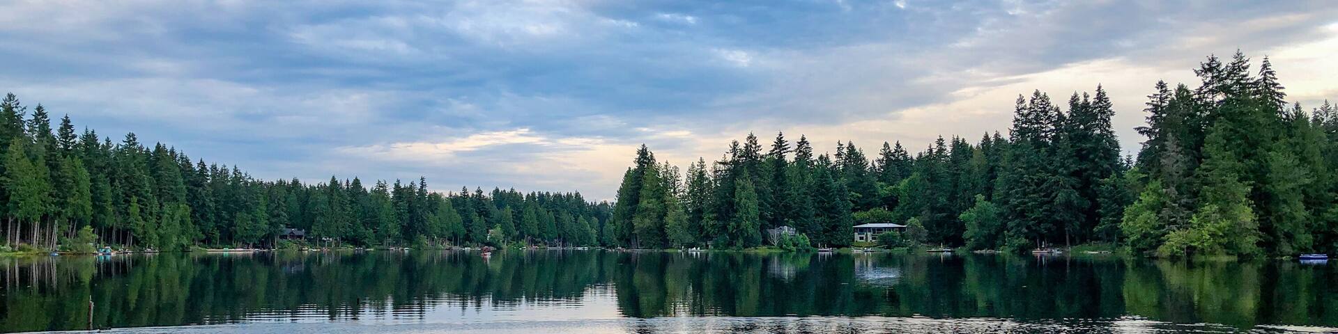 A calm lake surrounded by dense forest and a small house near the shore.
