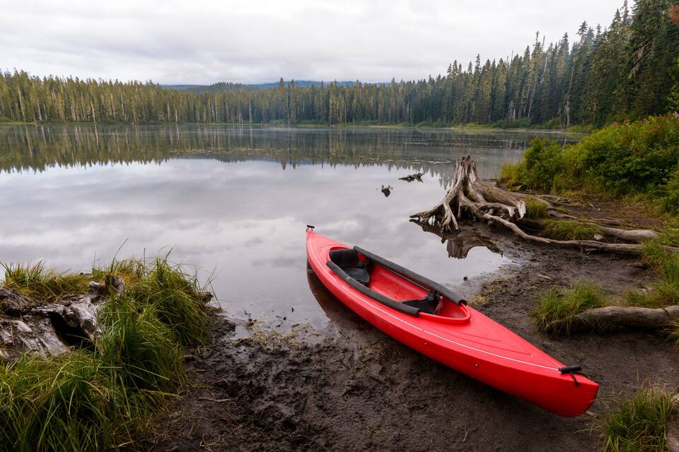 A red kayak on a muddy shore with a calm lake and dense forest in the background.