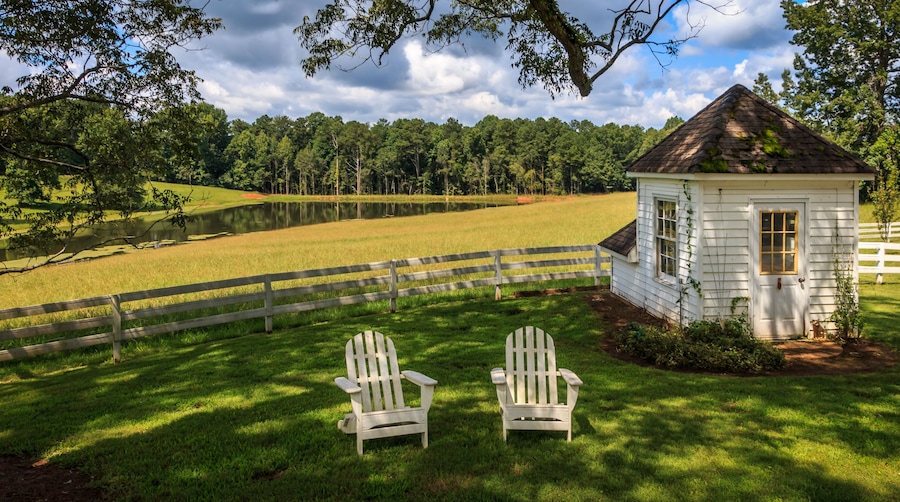 A white wooden cabin with a red roof, two Adirondack chairs, a wooden fence, and a pond surrounded by trees.