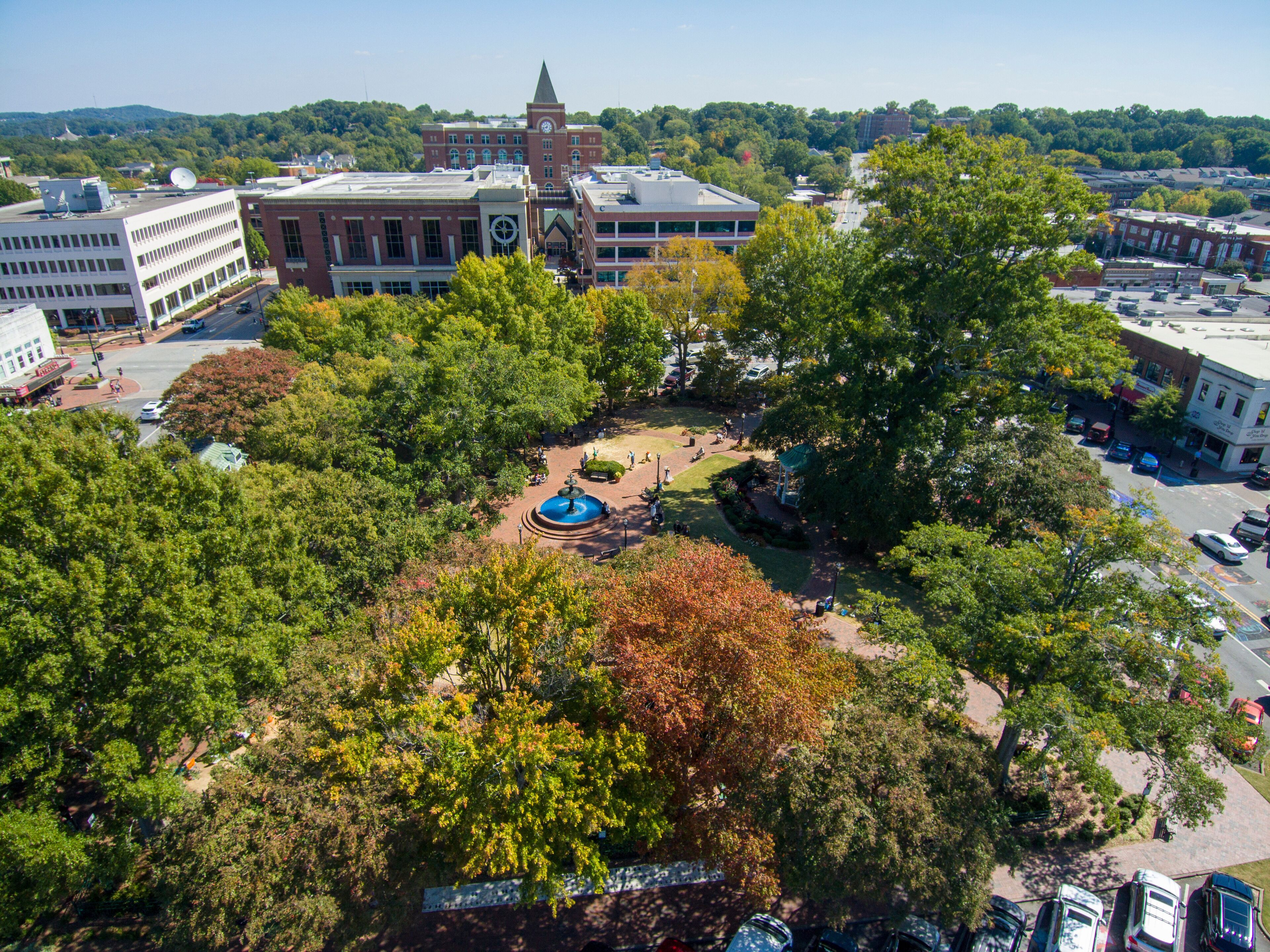 an aerial shot of a gorgeous autumn landscape at the Marietta Square with a water fountain surrounded by lush green trees and autumn colored trees with buildings and blue sky in Marietta Georgia USA