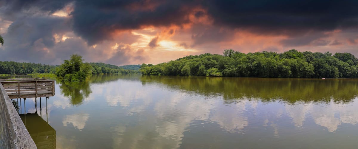 a panoramic shot of the still silky brown water of the Chattahoochee river with a long wooden boardwalk along the river and lush green trees with powerful clouds at Roswell Riverwalk in Georgia