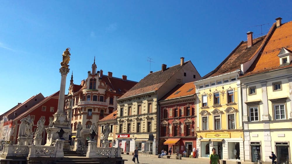 Ein historischer Platz mit kunstvollen Gebäuden, einem zentralen Brunnen und Fußgängern.