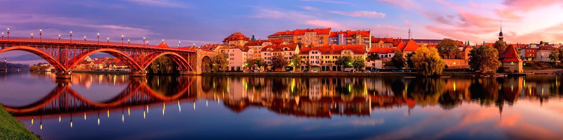 A red bridge over a calm river reflecting the surrounding buildings and sky.