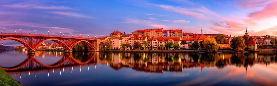 A red bridge over a calm river reflecting the surrounding buildings and sky.