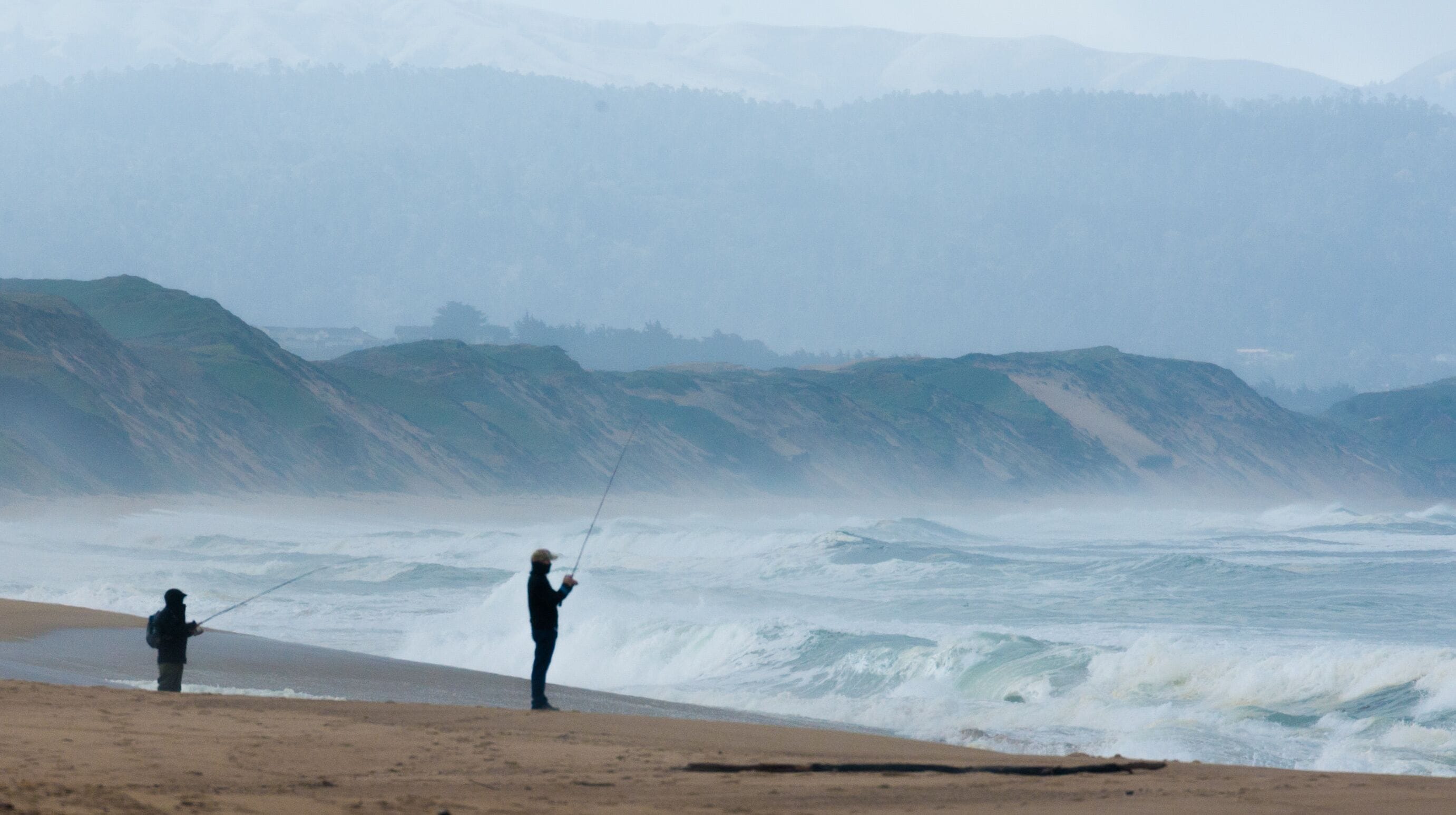 A beach with waves crashing onto the shore, a person fishing, and a hilly landscape in the background.