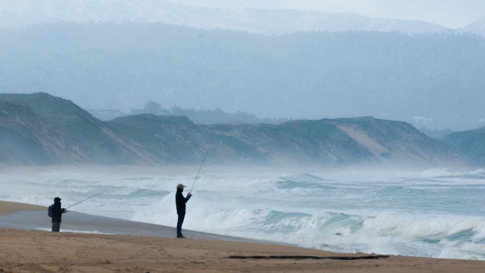 A beach with waves crashing onto the shore, a person fishing, and a hilly landscape in the background.
