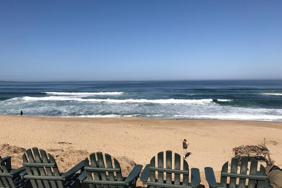 A beach with waves, a clear sky, and green Adirondack chairs.
