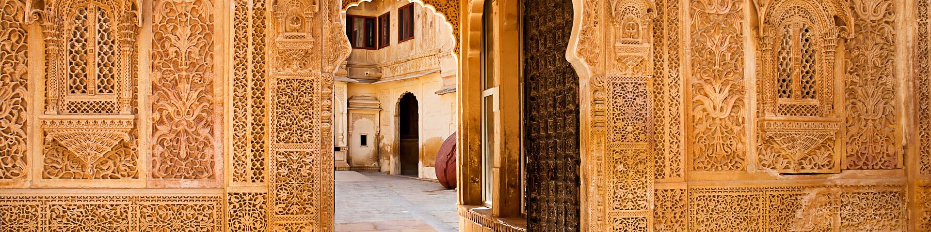 An ornate archway with intricate carvings, leading to a courtyard with a fountain and a building in the background.