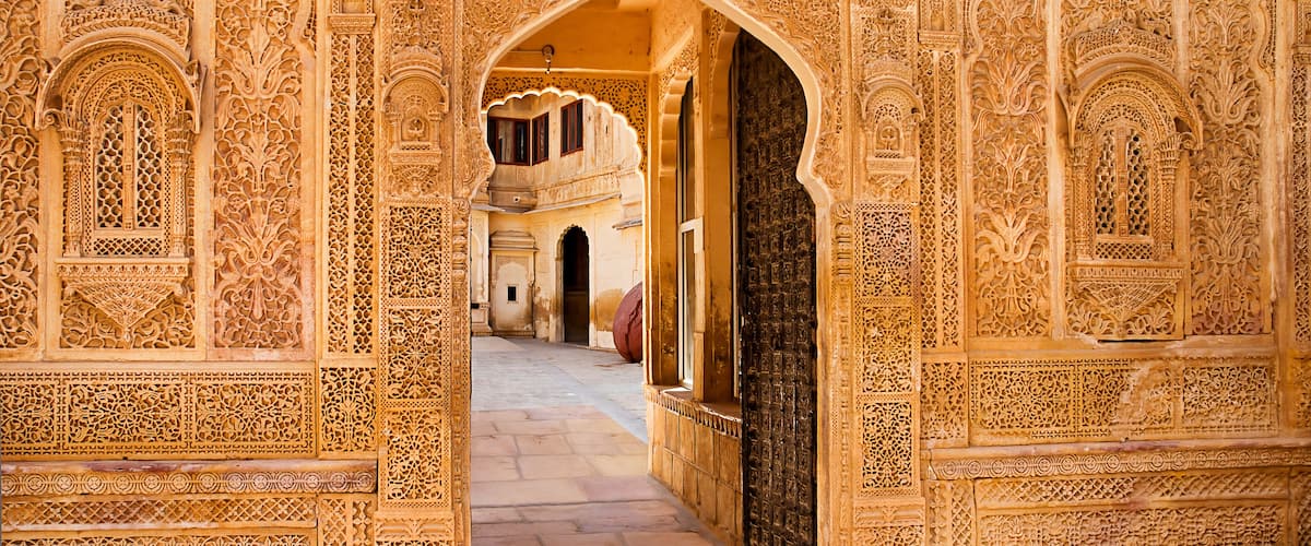 An ornate archway with intricate carvings, leading to a courtyard with a fountain and a building in the background.