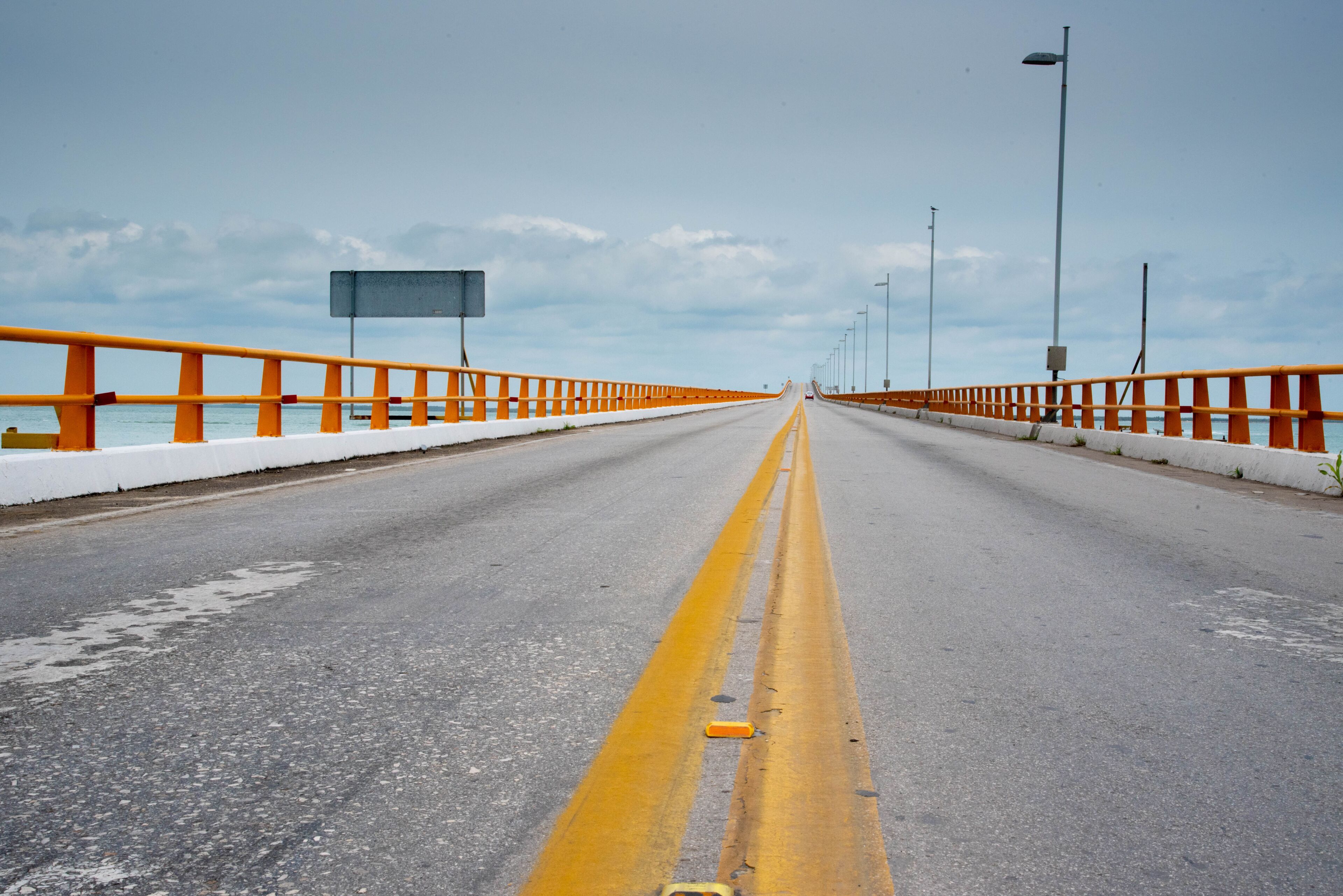 Un puente con barandillas anaranjadas, cielo despejado y una carretera con marcas amarillas en el centro.