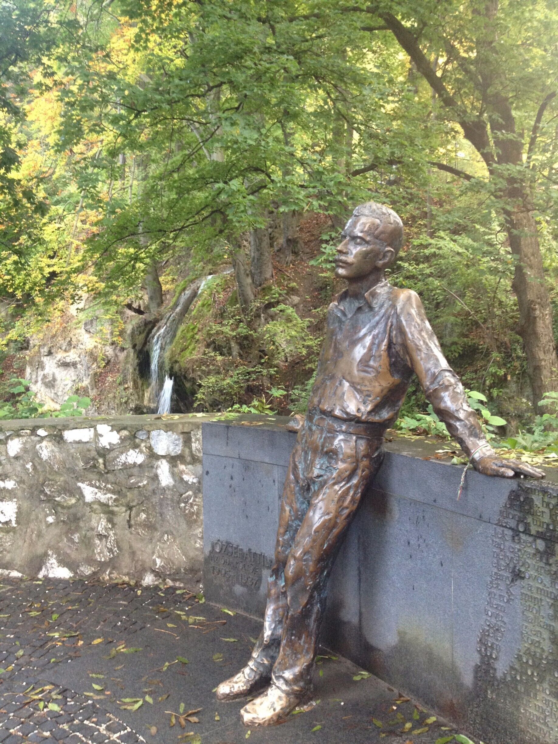 A bronze statue of a man leaning on a wall, surrounded by trees and a small waterfall.