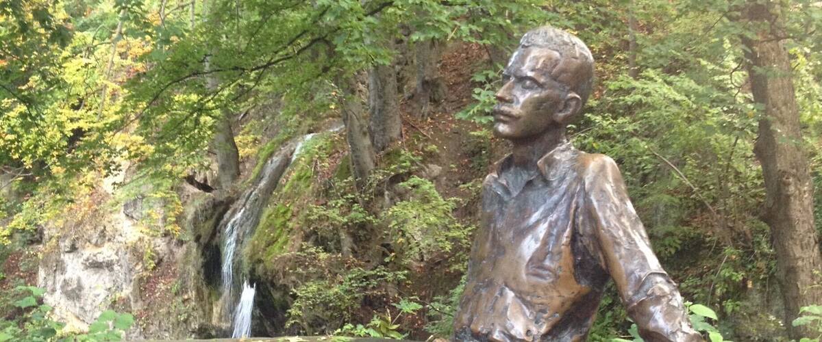 A bronze statue of a man leaning on a wall, surrounded by trees and a small waterfall.